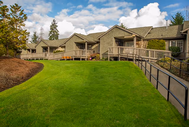Exterior view of a single-story senior living facility building with multiple units, each having a small porch and wheelchair-accessible ramps. The building is surrounded by a well-maintained green lawn, trees, and shrubs under a partly cloudy sky.