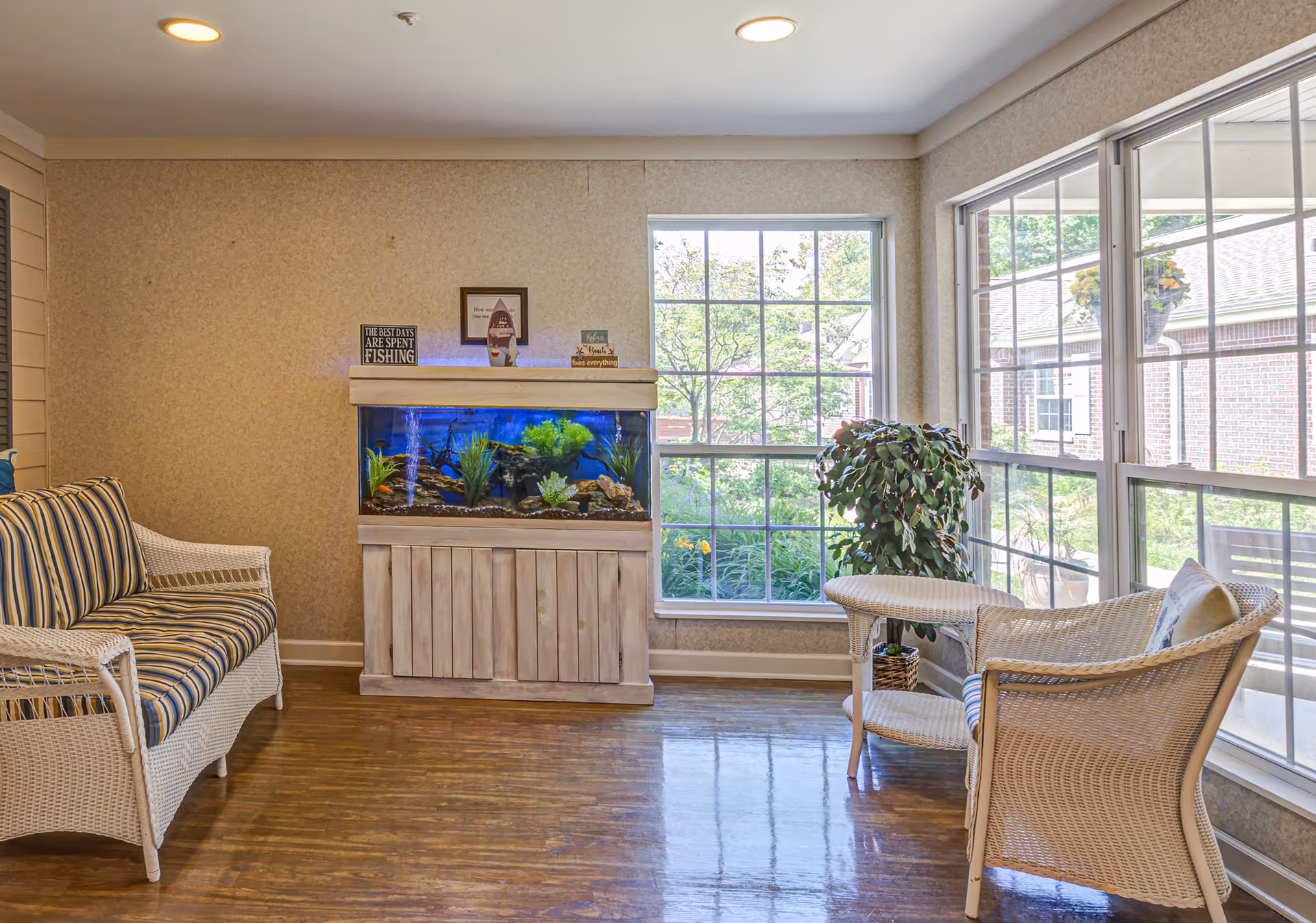 A cozy sitting area with wicker furniture including a striped cushioned loveseat and a chair with a small matching table. There is a large fish tank on a wooden stand against the wall, and large windows letting in natural light with a view of greenery outside. A potted plant is placed near the windows.