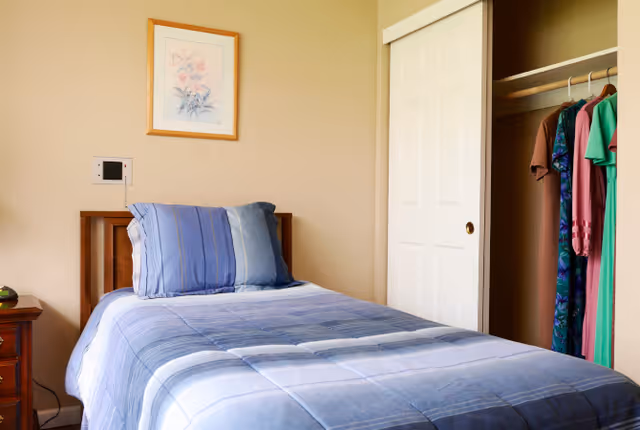 Sunlit small bedroom with a single bed dressed in blue linens, a nightstand, wall art, and an open closet with hanging clothes.
