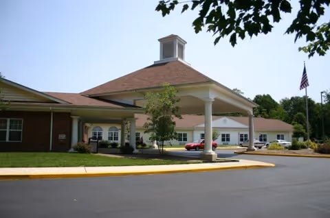 Exterior view of a single-story brick and white building with a covered entrance supported by columns. There are several cars parked in front, a small tree near the entrance, and an American flag on a flagpole to the right. The sky is clear and blue.