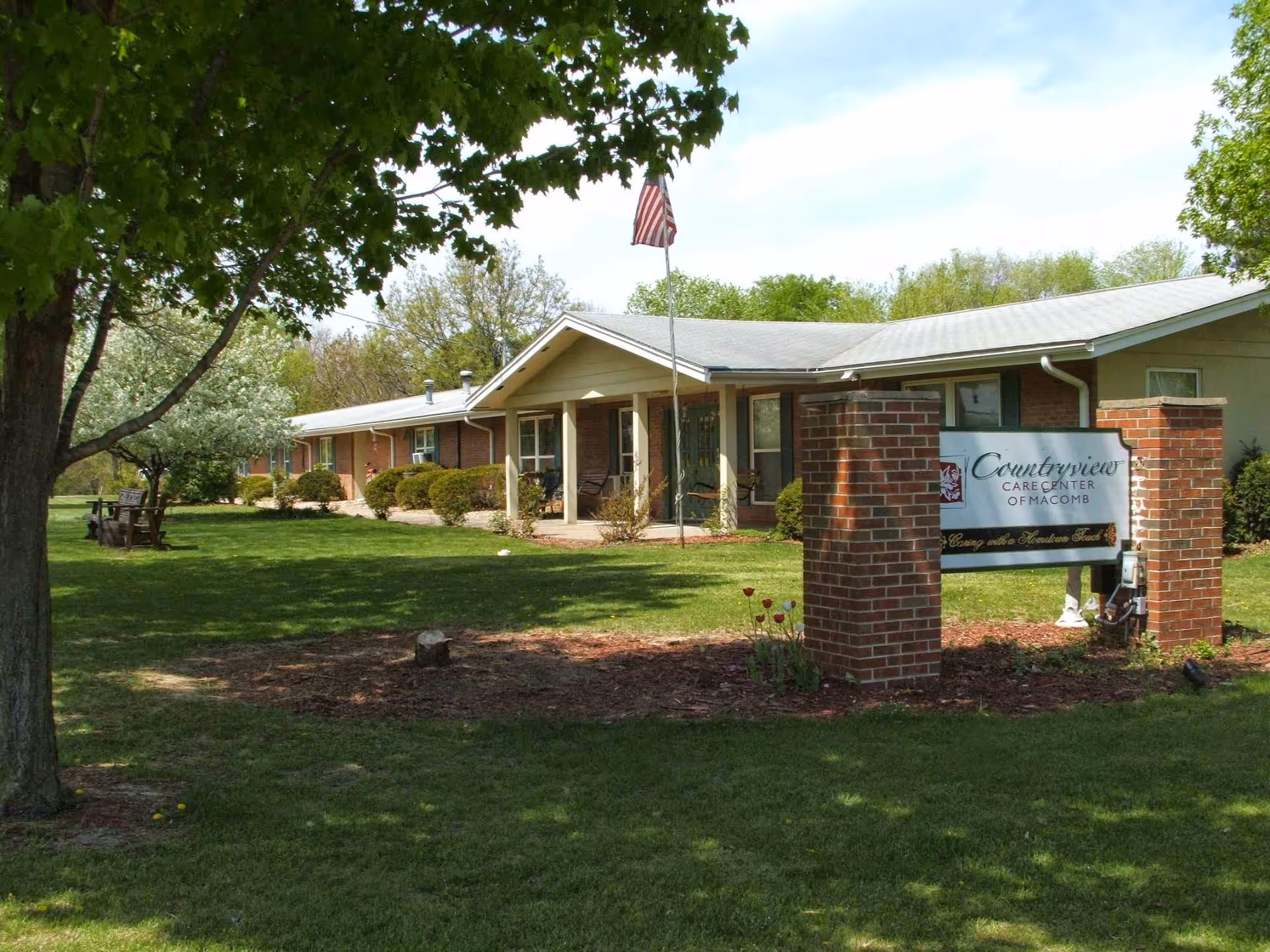Exterior view of Countryview Care Center of Macomb, a single-story brick building with a covered entrance and an American flag on a flagpole in front. The building is surrounded by green grass, trees, and shrubs, with a sign displaying the facility's name near the entrance.
