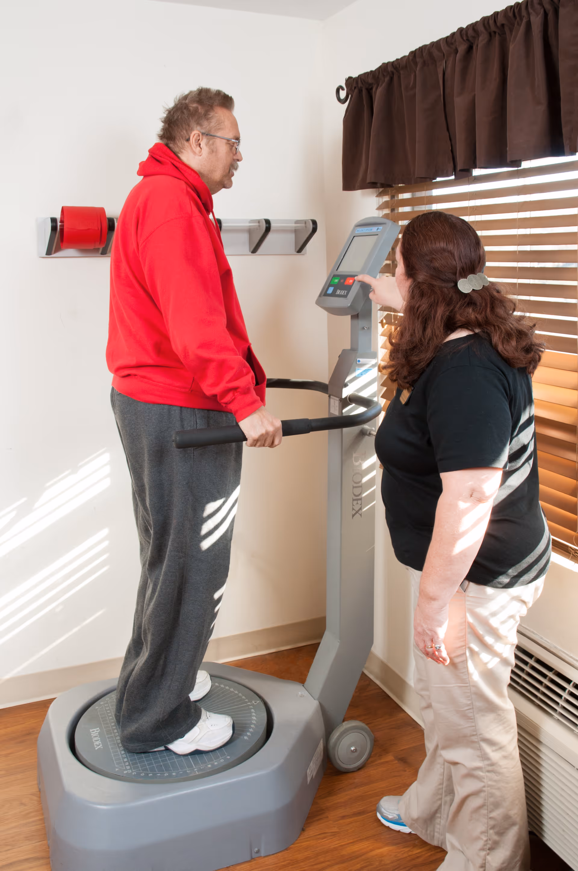 A man in a red hoodie and gray sweatpants stands on a vibration exercise machine while a woman in a black shirt and beige pants operates the machine's control panel in a room with wooden blinds and hardwood flooring.