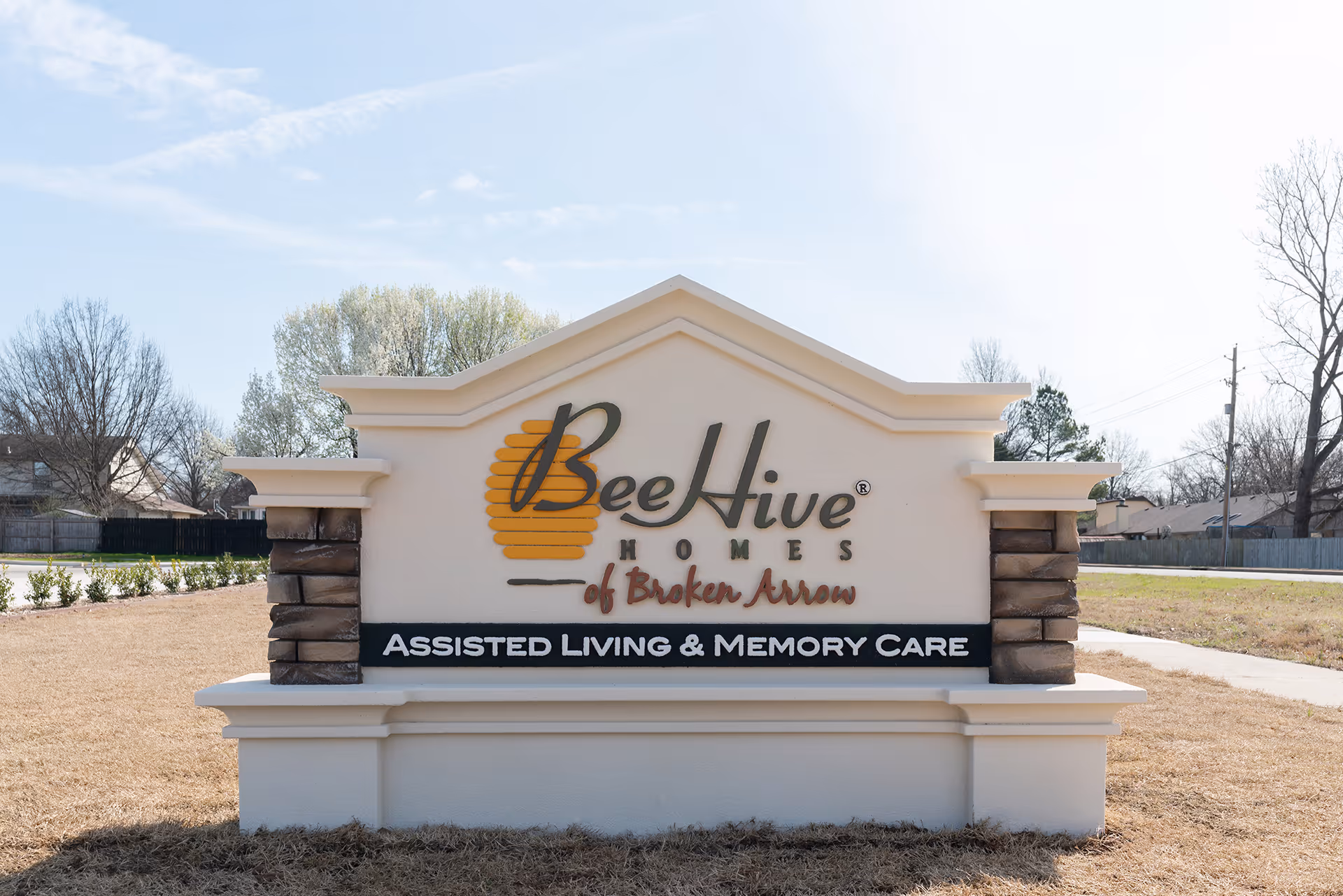 Outdoor sign for BeeHive Homes of Broken Arrow, an assisted living and memory care facility, set on a grassy area with trees and houses in the background under a partly cloudy sky.