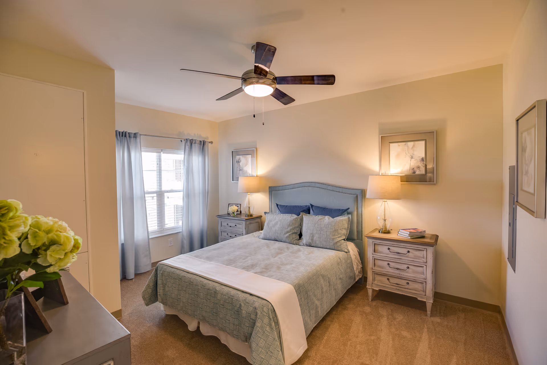 A cozy bedroom with a neatly made bed featuring a light blue upholstered headboard, blue and gray pillows, and a light blue bedspread. There are two matching nightstands on either side of the bed, each with a lamp and decorative items. A window with light blue curtains allows natural light into the room. The walls are painted a soft beige, and a ceiling fan with dark blades is mounted above the bed. A dresser with a vase of green flowers is partially visible in the foreground.