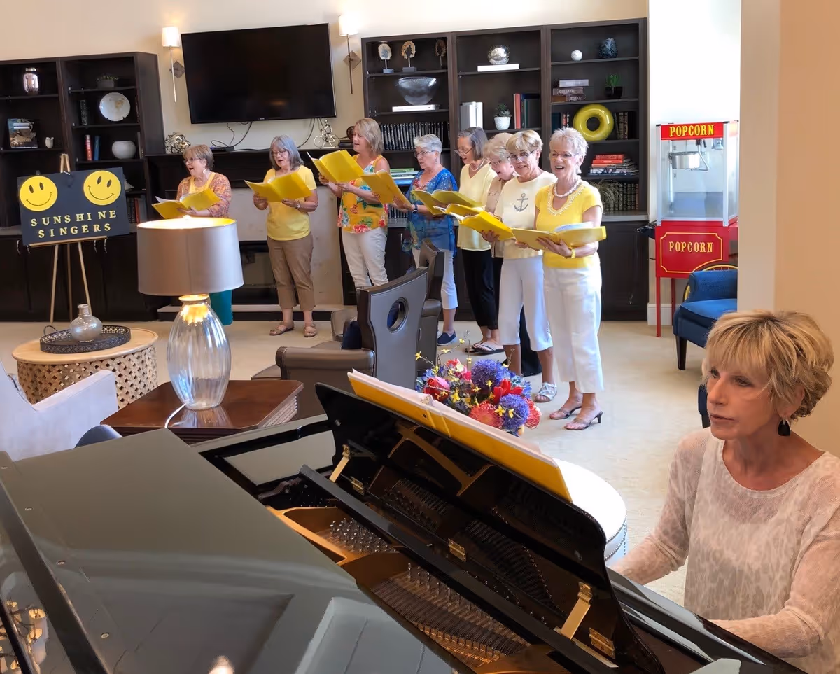 A group of elderly women standing in a line singing from yellow songbooks in a living room setting. A woman is playing a grand piano in the foreground. Behind the singers is a sign that reads 'Sunshine Singers' with two smiley faces, a large flat-screen TV mounted on the wall, bookshelves, and a popcorn machine. The room is warmly lit and decorated with flowers and comfortable furniture.