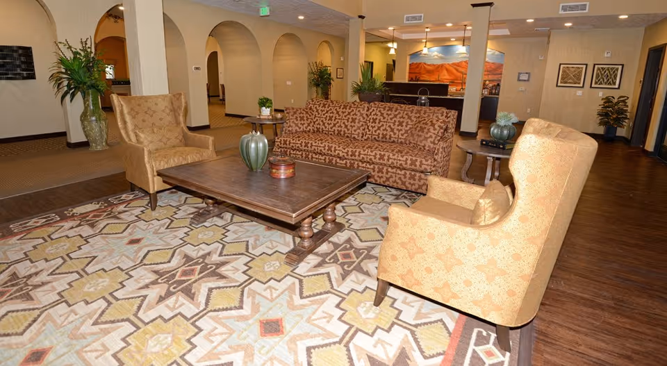 A cozy living room area in Jaidon Manor assisted living featuring a patterned sofa and two matching armchairs arranged around a wooden coffee table on a geometric patterned rug. The room has warm lighting, decorative plants, framed artwork on the walls, and arched doorways leading to other parts of the facility.