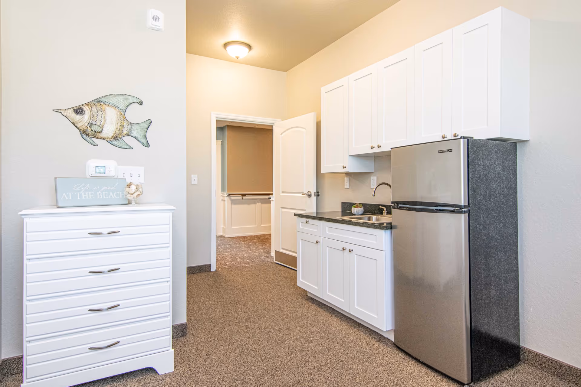A small kitchenette area with white cabinets, a stainless steel refrigerator, and a sink. To the left, there is a white chest of drawers with a decorative fish wall hanging above it and a sign that reads 'Life is good AT THE BEACH'. The room has beige walls and carpeted flooring, with an open door leading to another room.