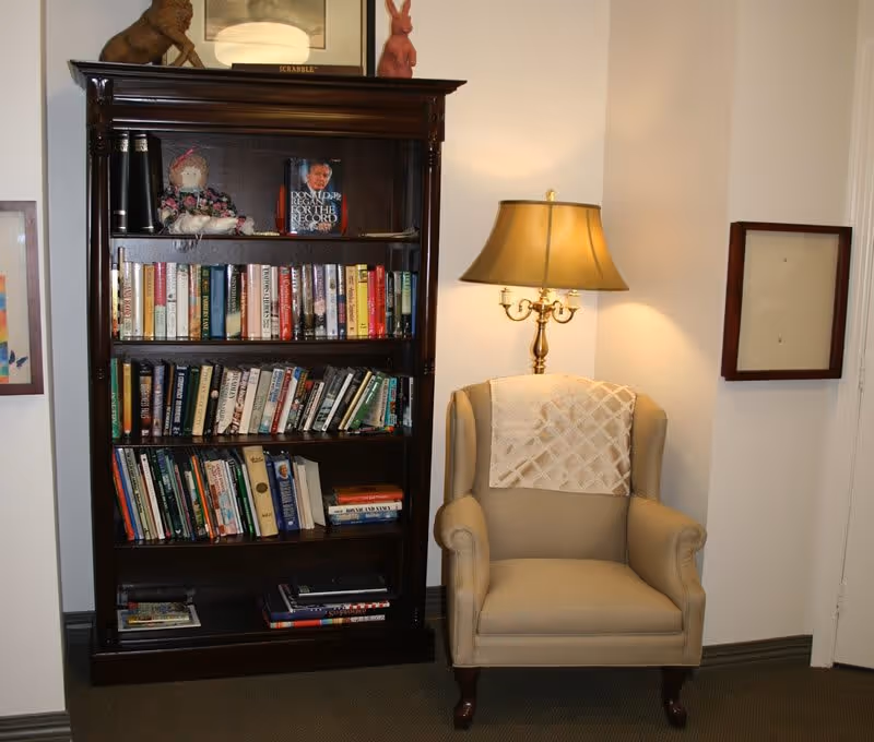 A cozy reading corner with a tall dark wood bookshelf filled with books beside an upholstered armchair and a lit floor lamp.