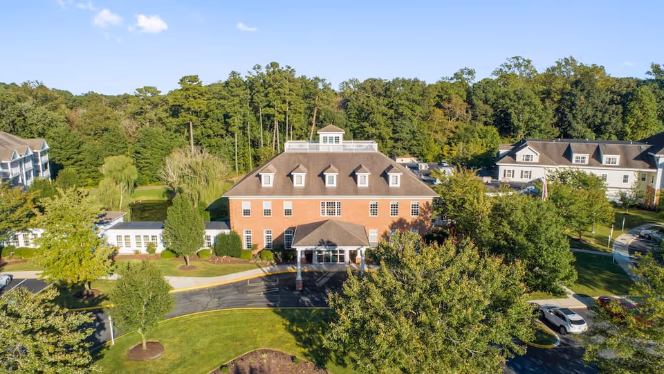 Aerial view of a brick senior living facility building with a covered front entrance, surrounding trees, lawns, and parked cars.