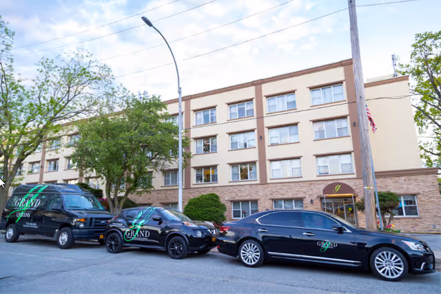 Front exterior of a four-story senior living facility with an entrance awning and three black cars and a van parked in front.