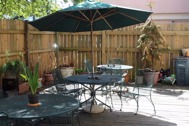 Outdoor patio area with metal tables and chairs, one table has a large green umbrella. Several potted plants and a wooden fence surround the space, with sunlight casting shadows on the wooden deck floor.