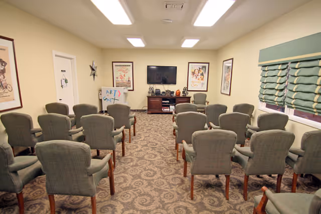 A small meeting or activity room with multiple rows of green upholstered chairs facing a wall-mounted flat screen TV. The room has beige walls, patterned carpet, framed posters on the walls, a window with green blinds, and a small cabinet under the TV holding various items. A whiteboard with colorful writing is visible on the left side of the room.