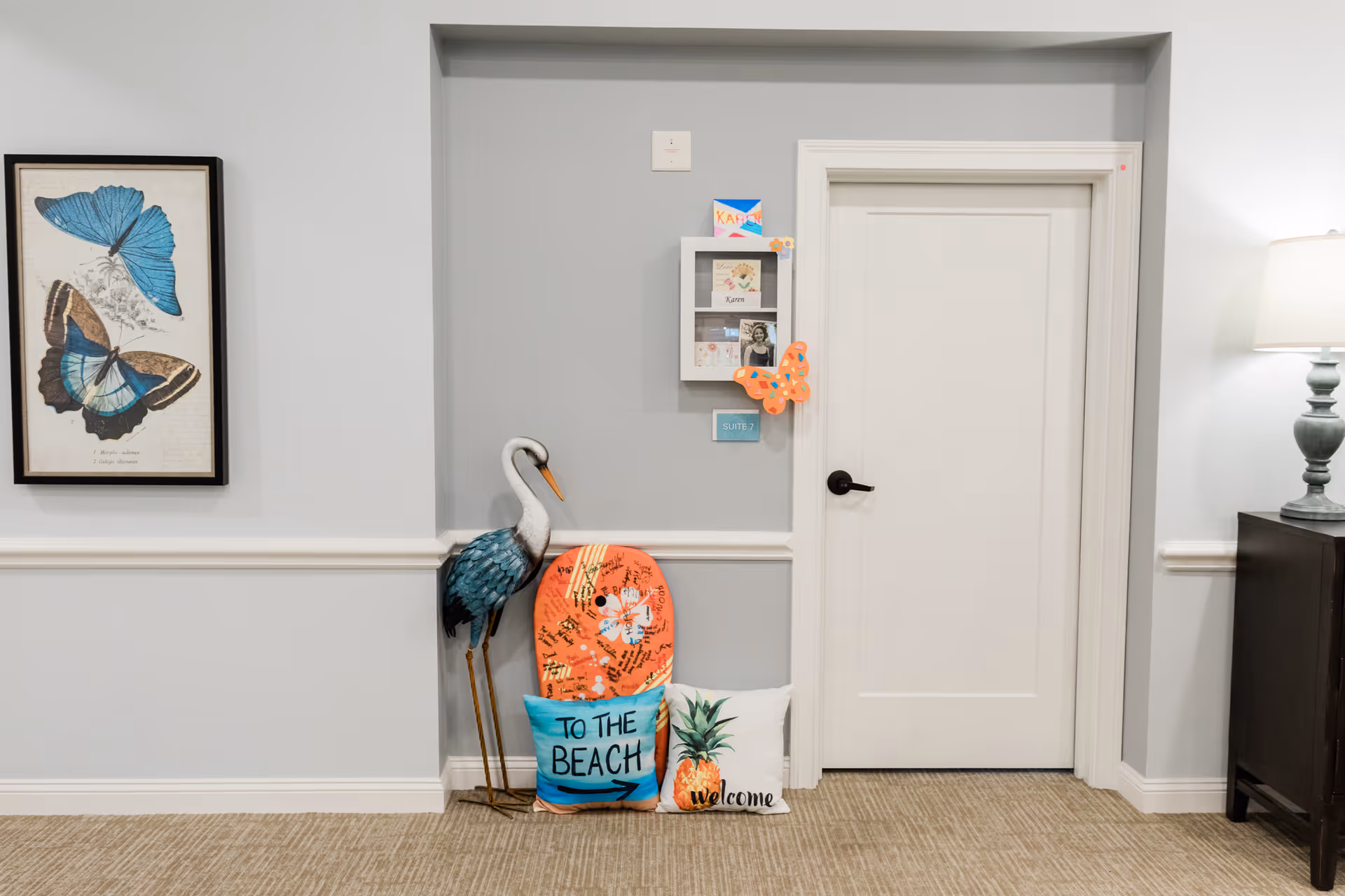 Interior hallway of a memory care facility with a closed white door labeled Suite 7. Next to the door is a decorative metal crane statue, a colorful boogie board, and two decorative pillows, one with the text 'TO THE BEACH' and the other with a pineapple and the word 'welcome'. A framed butterfly artwork hangs on the left wall, and a lamp on a dark cabinet is visible on the right.