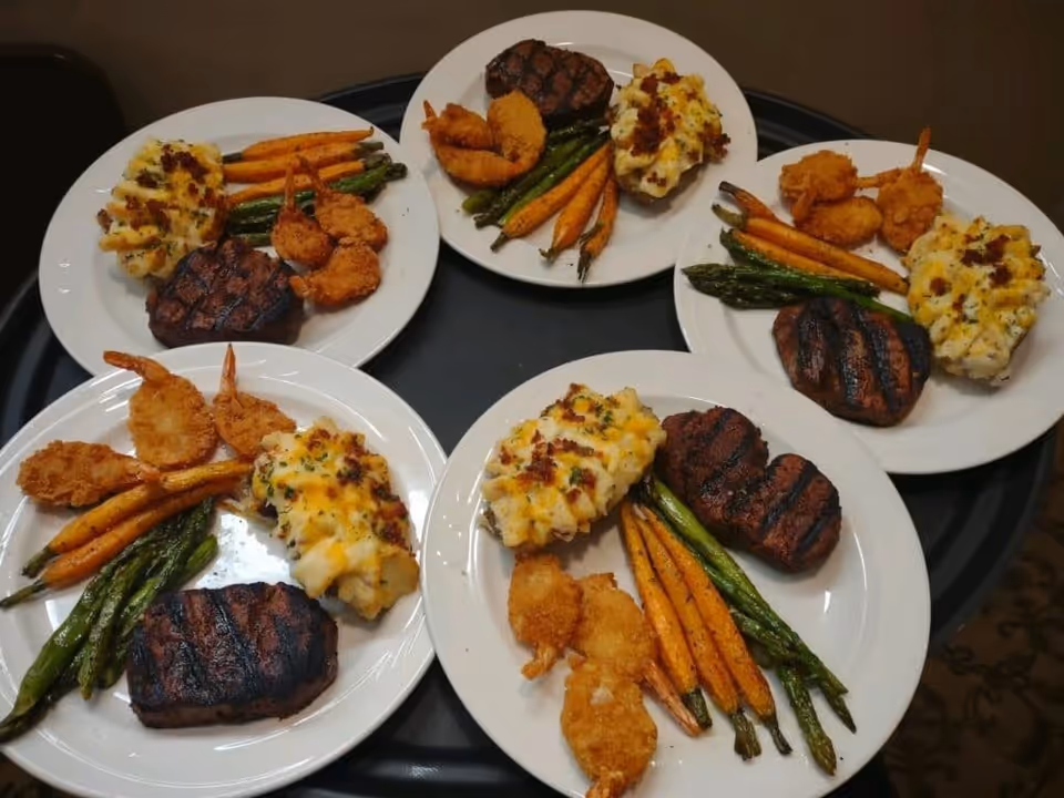 Five plates of food on a black tray, each plate containing a grilled steak, breaded fried shrimp, roasted carrots and asparagus, and a serving of loaded mashed potatoes topped with cheese and bacon bits.