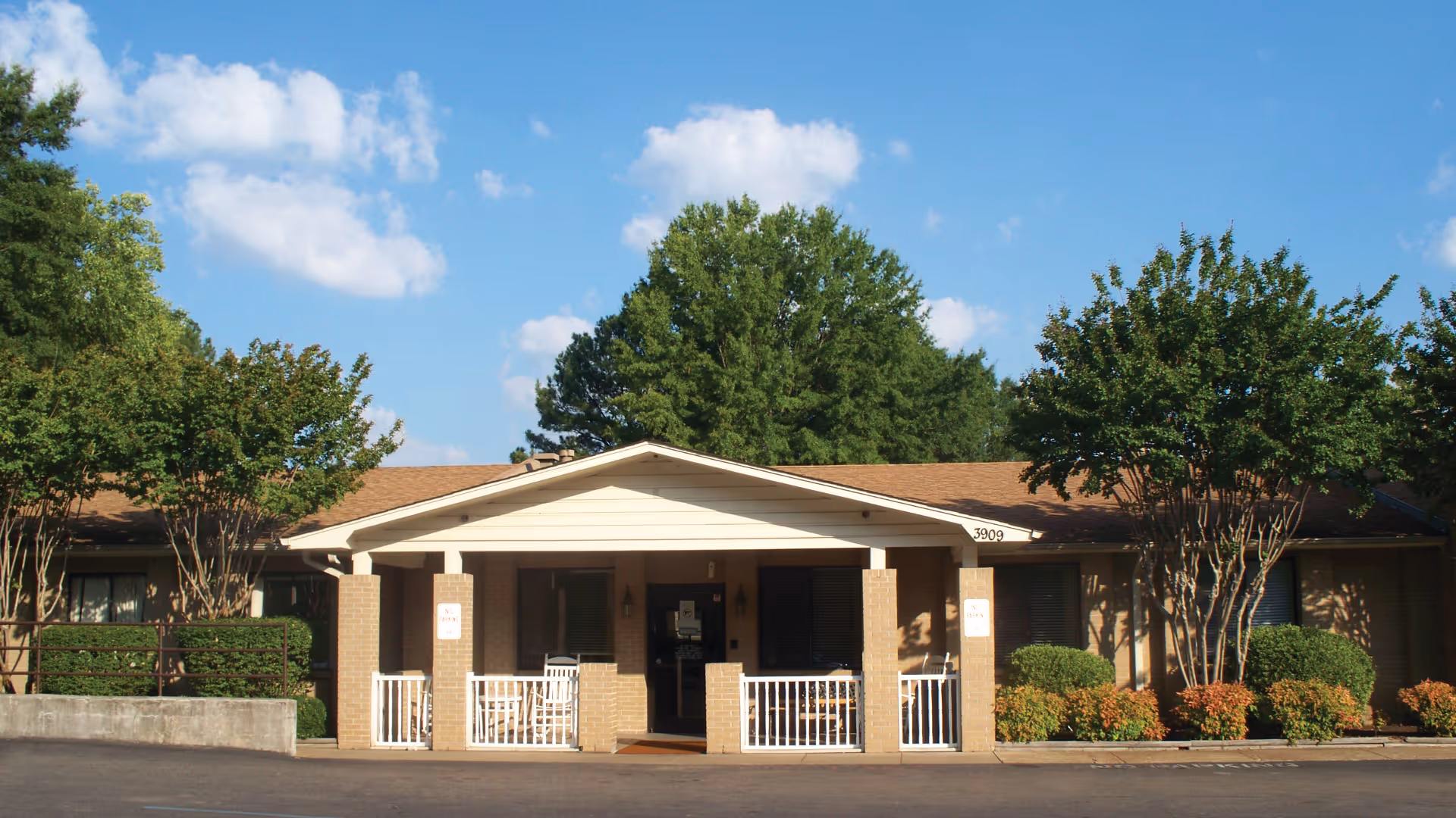Front exterior view of Spring Gate Rehabilitation and Health Care Center showing a single-story building with a covered entrance, white railings, brick pillars, surrounded by trees and shrubs under a blue sky with some clouds.