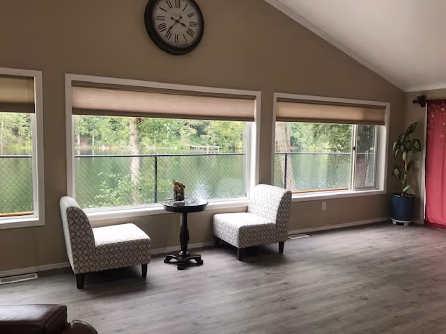 A bright living room with large windows overlooking a green outdoor area with a lake. The room has two patterned chairs facing each other with a small round table between them. A large clock is mounted on the wall above the windows. There is a potted plant in the corner near a red curtain.