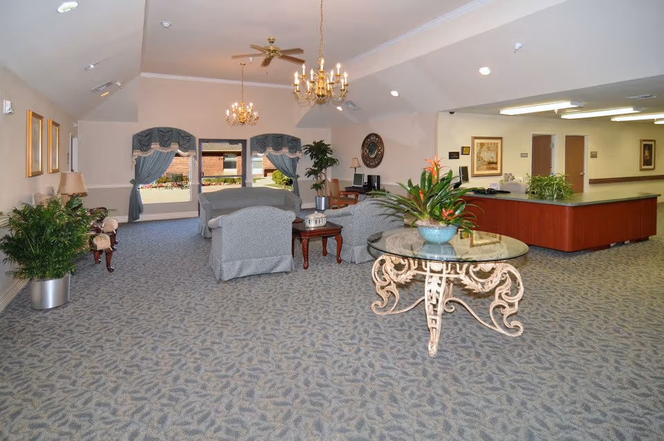 A spacious and well-lit common area in a senior living facility featuring a seating arrangement with upholstered chairs and a sofa around a wooden coffee table. There is a decorative round glass table with a potted plant in the foreground. The room has carpeted floors, two chandeliers, ceiling fans, and large windows with blue curtains. A reception desk is visible in the background along with framed artwork on the walls and additional plants.