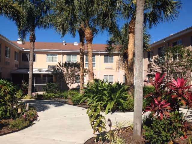 Exterior view of a two-story assisted living facility with peach-colored walls and a red-tiled roof, surrounded by tall palm trees and lush tropical plants under a clear blue sky.