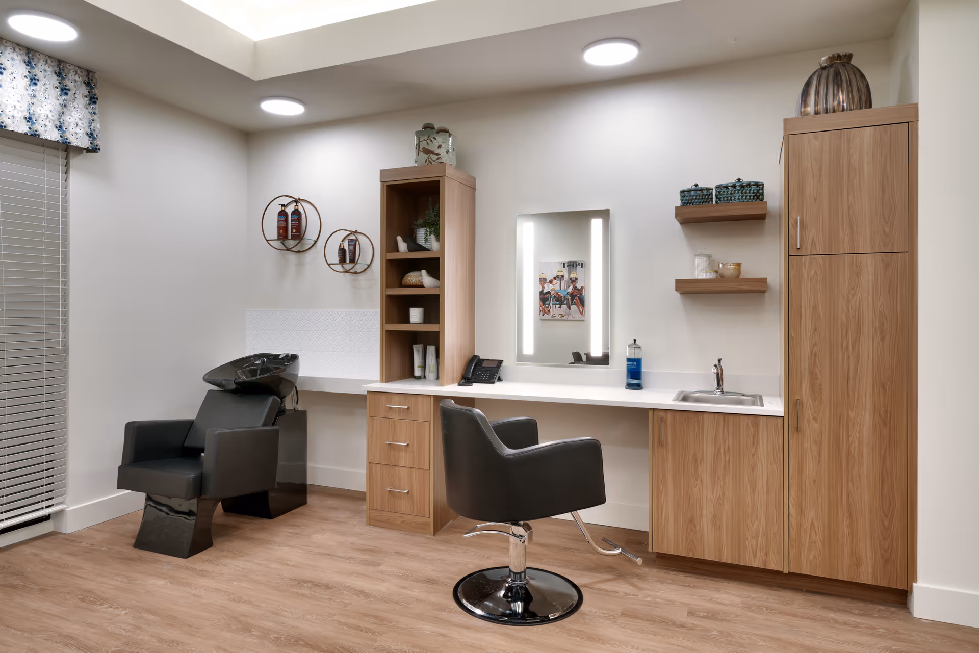 A modern salon area in a senior living facility featuring a black salon chair in front of a mirror with built-in lights, a black hair washing station with a reclining chair, wooden cabinetry with shelves holding decorative items and hair products, a small sink, and light wood flooring.
