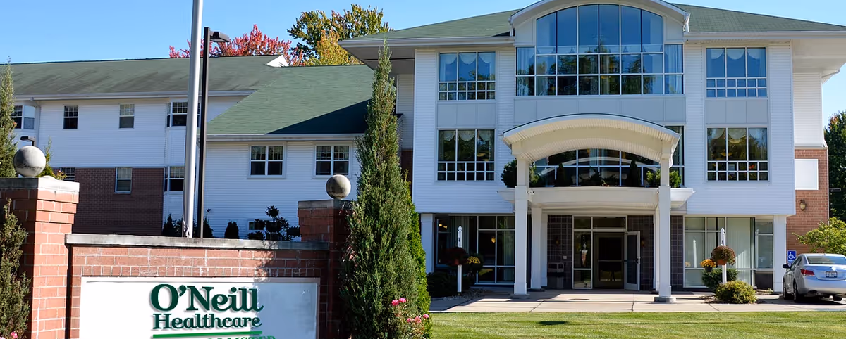 Front exterior view of O'Neill Healthcare North Olmsted building with white siding, large windows, a green roof, and a covered entrance. There is a brick sign in the foreground with the facility name and some landscaping including trees and bushes.