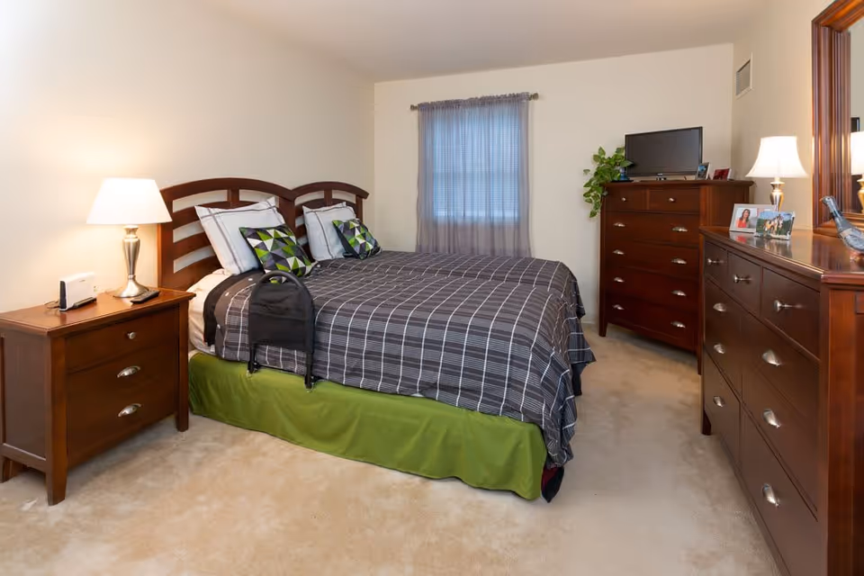 A neatly arranged bedroom with a double bed covered in a gray checkered bedspread and green bed skirt. The bed has four pillows, two with geometric patterns. There is a wooden nightstand with a lamp and a small device on it to the left of the bed. A window with sheer curtains is centered on the back wall. To the right, there are two wooden dressers, one with a TV and a plant on top, and the other with a lamp and framed photos. The room has beige carpet and cream-colored walls.