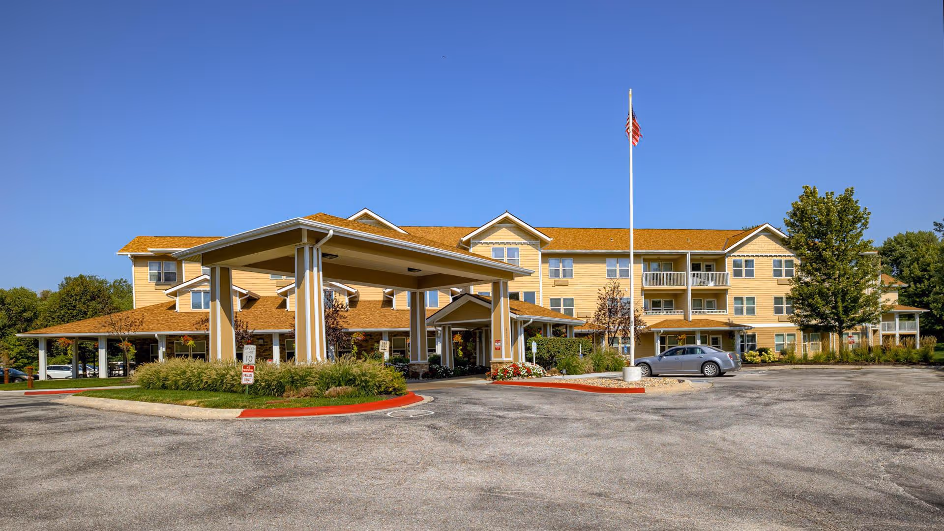 Exterior view of a three-story senior living facility building with a covered entrance driveway, surrounded by landscaping and a parking area. An American flag is flying on a flagpole in front of the building under a clear blue sky.