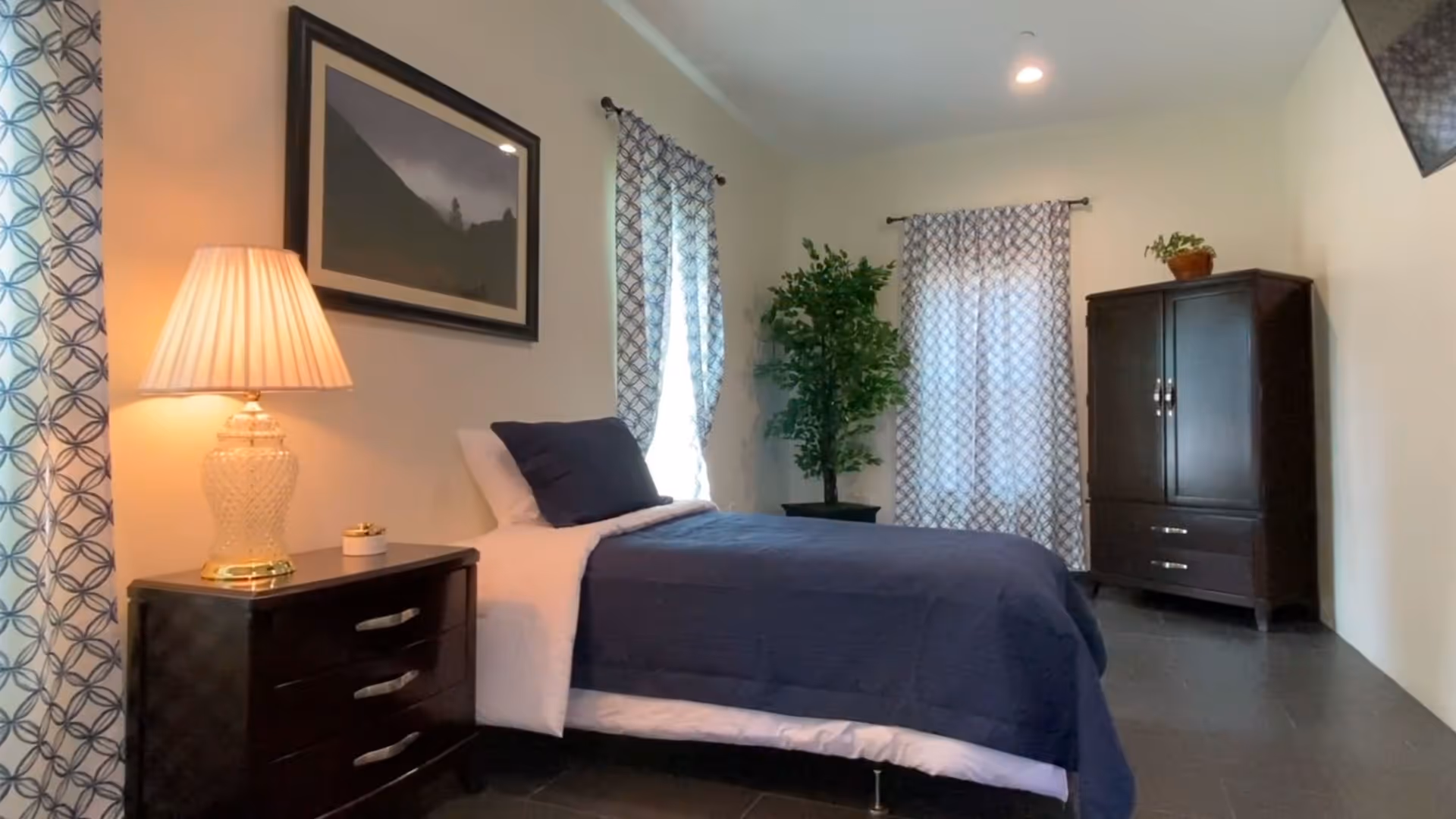 A neatly furnished bedroom with a single bed, bedside table and lamp, wardrobe, potted plant, and patterned curtains.