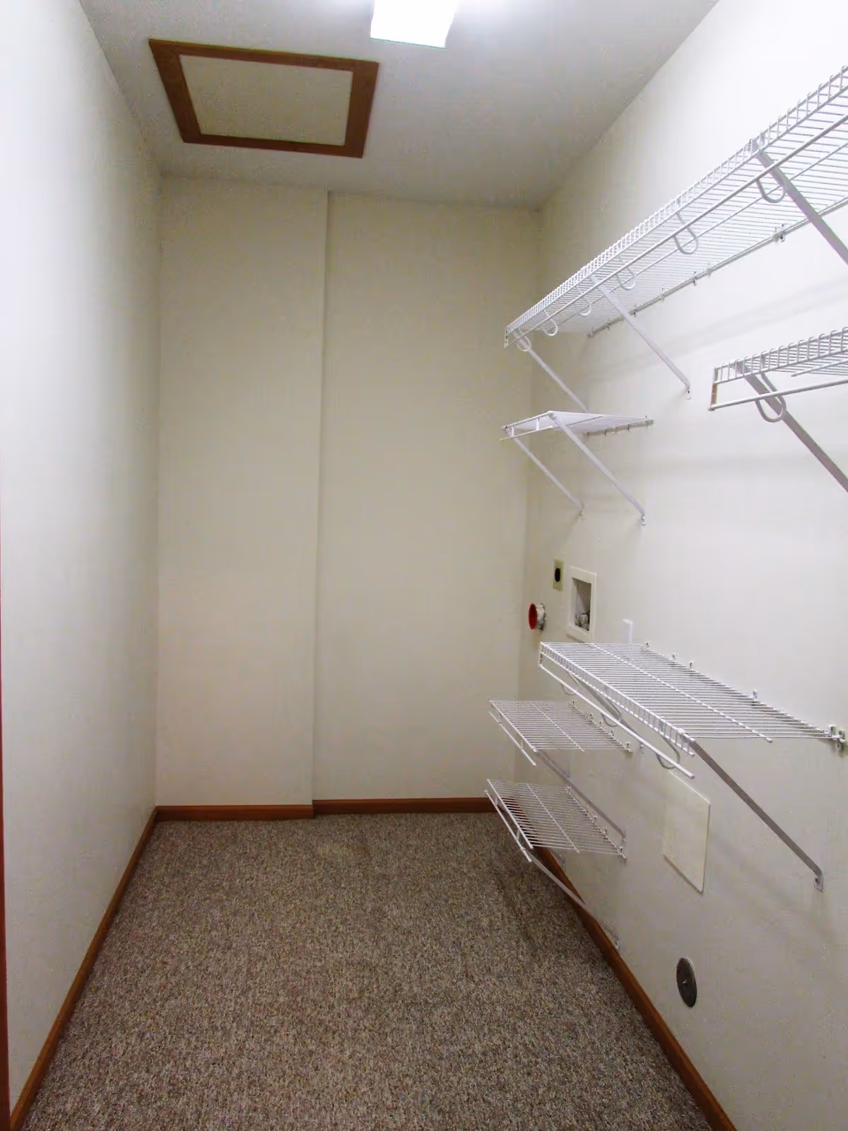 Empty laundry room with beige walls, carpeted floor, and multiple white wire shelves mounted on the right wall. There is a ceiling light and a small access panel on the ceiling.
