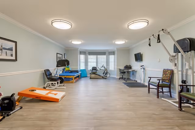 A spacious exercise room with light-colored walls and wood flooring. The room contains various fitness equipment including a stationary bike, elliptical machine, weight machine, exercise mats, and a cornhole game board. There are chairs along the walls and a wall-mounted TV. Large windows with blinds allow natural light into the room.