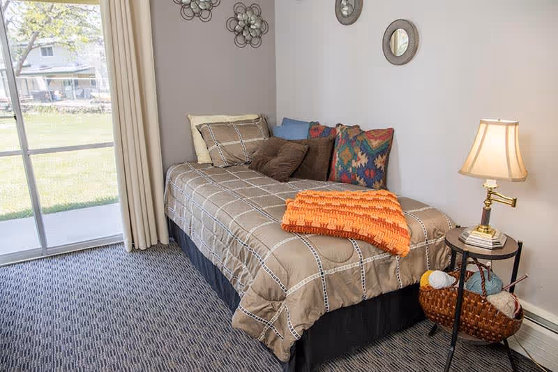 A small bedroom with a single bed covered in a beige checkered comforter and multiple pillows, including colorful patterned ones. An orange knitted blanket is folded at the foot of the bed. Next to the bed is a round side table with a lamp, some books, and a basket containing yarn and knitting needles. A sliding glass door with beige curtains leads to an outdoor grassy area. The walls are decorated with three small round mirrors and metal flower wall art.