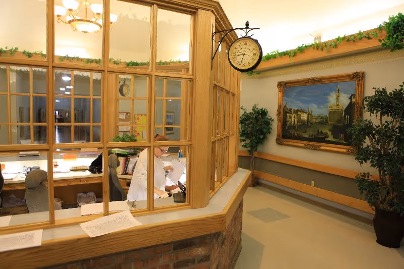 Reception area inside a senior living facility with a wooden framed glass partition. A staff member is working behind the counter. The area is decorated with potted plants, a large framed painting on the wall, and a vintage-style clock hanging from the ceiling.