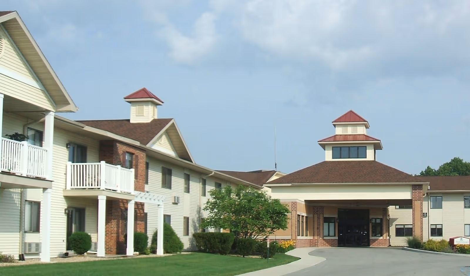 Exterior view of Pines Village Retirement Communities building with a driveway leading to the entrance, featuring beige siding, brick accents, balconies, and a well-maintained lawn with shrubs and trees under a partly cloudy sky.