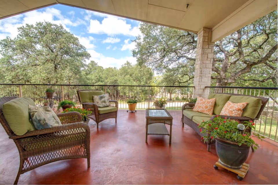 Covered outdoor patio area with wicker furniture including two armchairs and a loveseat with green cushions and decorative pillows. A glass-top wicker coffee table is in the center. Several potted plants are placed around the patio. The patio overlooks a green landscape with large trees under a partly cloudy sky.