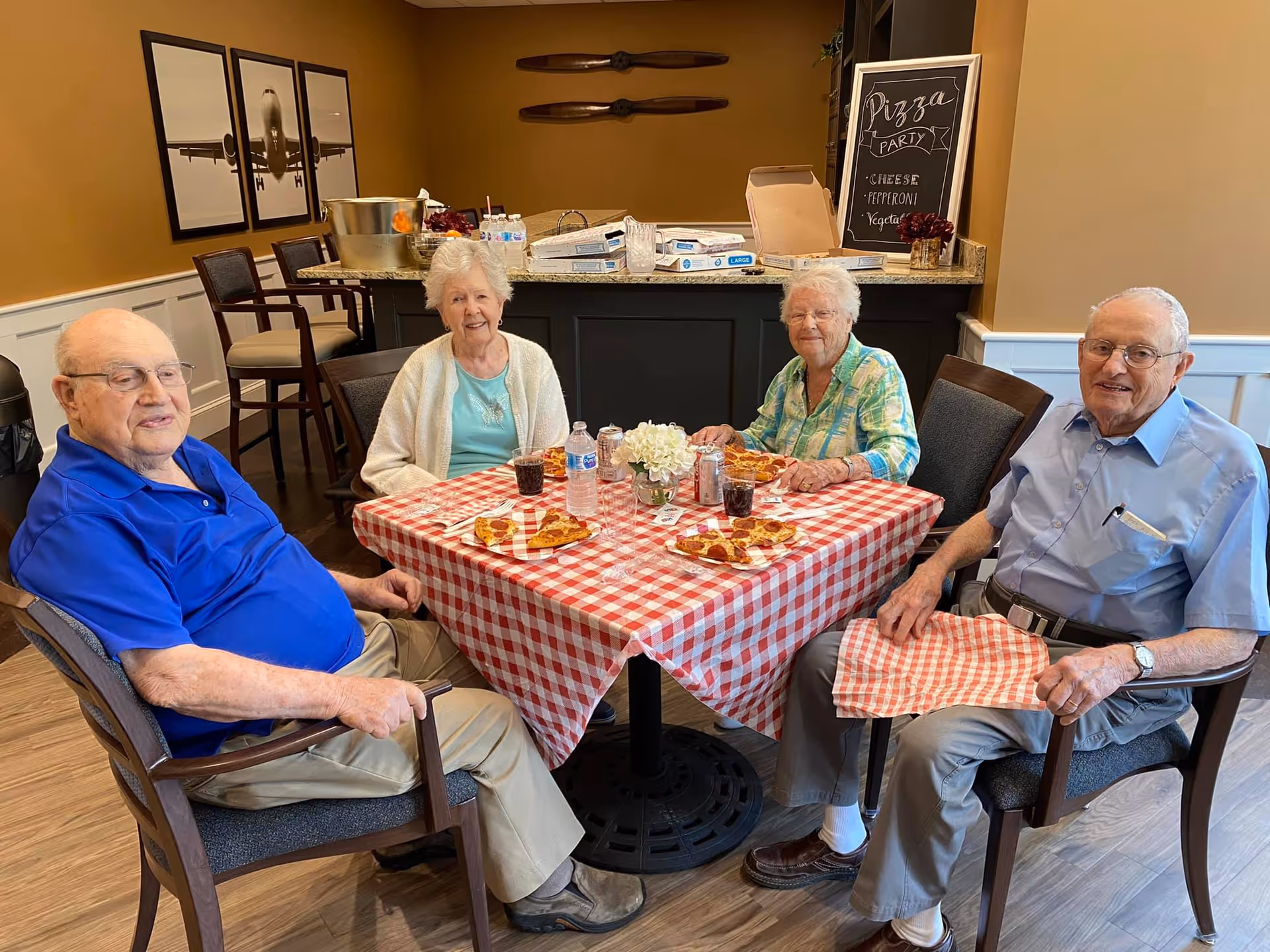 Four elderly people sitting around a table with a red and white checkered tablecloth, enjoying pizza and drinks in a cozy dining area. Behind them is a counter with pizza boxes and a chalkboard sign that reads 'Pizza Party' with cheese, pepperoni, and vegetable options listed.