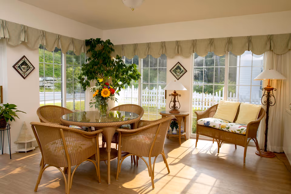 A bright and cozy sunroom with large windows and a view of a white picket fence outside. The room features a round glass-top wicker table surrounded by four wicker chairs with cushions. There is a wicker loveseat with floral cushions and two yellow pillows. Two decorative table lamps with white shades sit on wooden side tables. A large green potted plant and a vase with colorful flowers are also visible. The windows have green checkered valances.