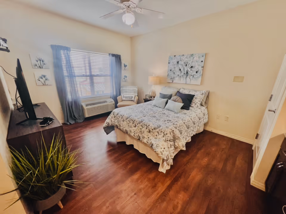 A cozy bedroom with a double bed covered in floral bedding and multiple pillows. There is a nightstand with a lamp and a small chair with a cushion near a window with blue curtains. A flat-screen TV is mounted on a stand opposite the bed, and a potted plant is placed on the floor. The room has wooden flooring and light-colored walls with floral artwork above the bed.