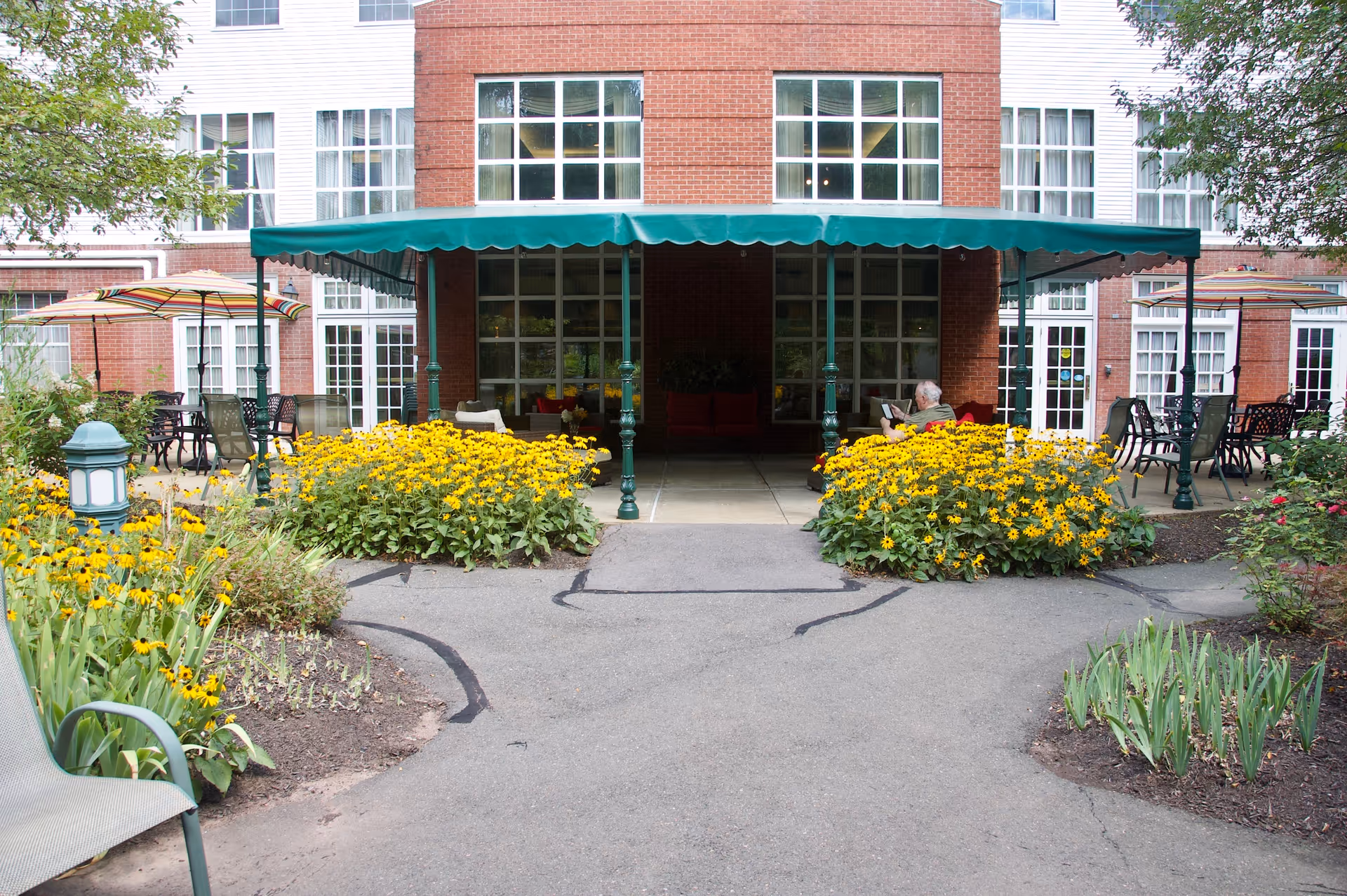 Outdoor patio area of a senior living facility with yellow flowers and green plants on either side of a paved walkway leading to a covered seating area with green canopy. There are tables with umbrellas and chairs on both sides, and a person is sitting and reading under the canopy. The building has large windows and red brick walls.