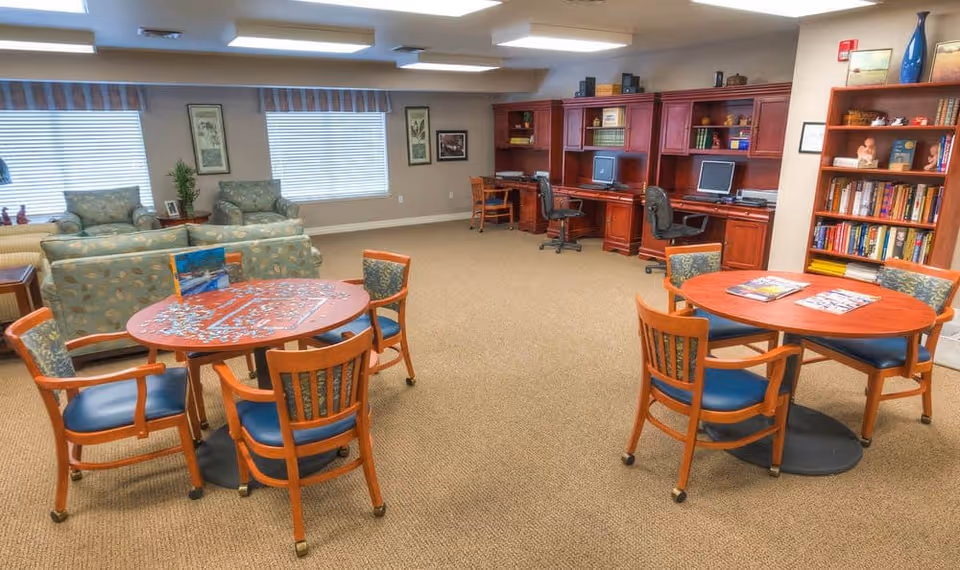 A senior living facility common area with two round wooden tables surrounded by chairs, a puzzle in progress on one table, and magazines on the other. In the background, there are two armchairs and a sofa near windows with blinds, and a wooden built-in desk area with computers and office chairs. A bookshelf filled with books and decorative items is also visible.