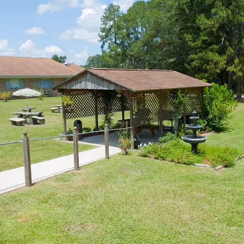 Outdoor courtyard with a wooden lattice gazebo, picnic tables, a small fountain, and a single-story building in the background.
