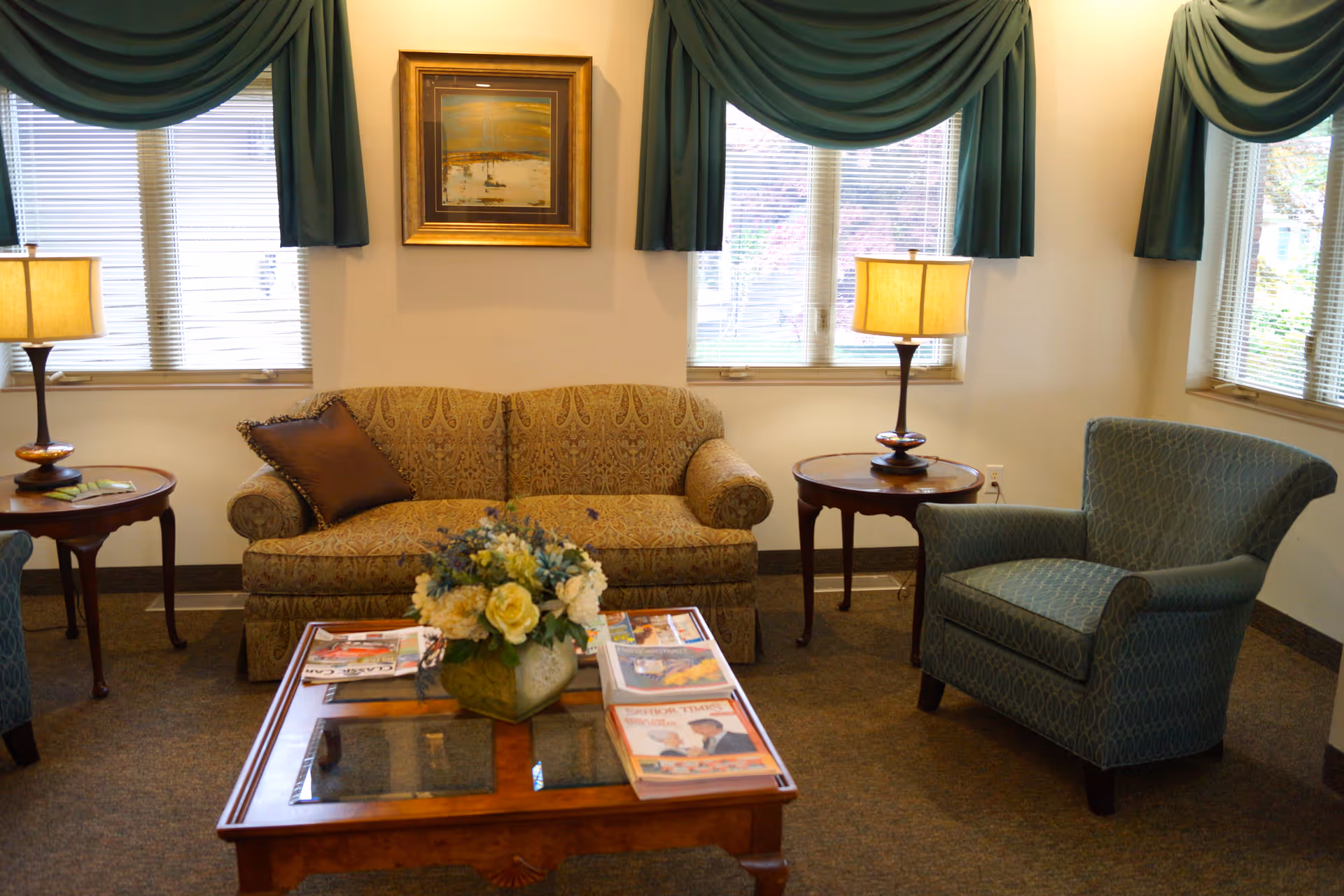 A cozy living room area with a patterned beige sofa, a blue armchair, two wooden side tables each with a lamp, a wooden coffee table with a glass top holding a flower arrangement and magazines, and three windows with green drapes and blinds. A framed painting hangs on the wall above the sofa.