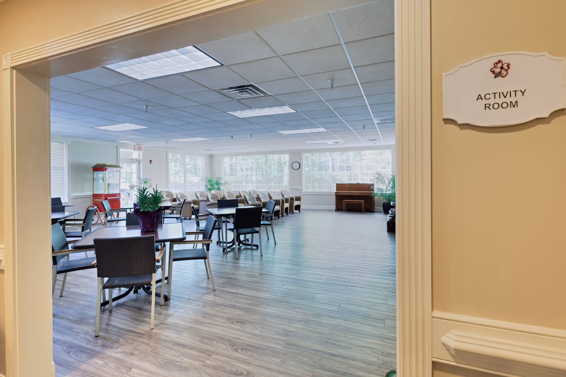 View through a doorway into a spacious activity room with multiple tables and chairs arranged for group activities. The room has large windows letting in natural light, a popcorn machine in the corner, a piano against the far wall, and some potted plants for decoration. A sign on the wall next to the doorway reads 'Activity Room'.