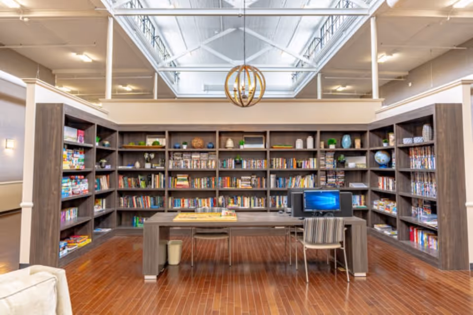 A spacious library or reading room with wooden bookshelves filled with books, board games, and decorative items. A large wooden table with a computer and chairs is positioned in the center of the room. The room has a high ceiling with a skylight and a modern circular chandelier hanging from it. The floor is made of polished wood.