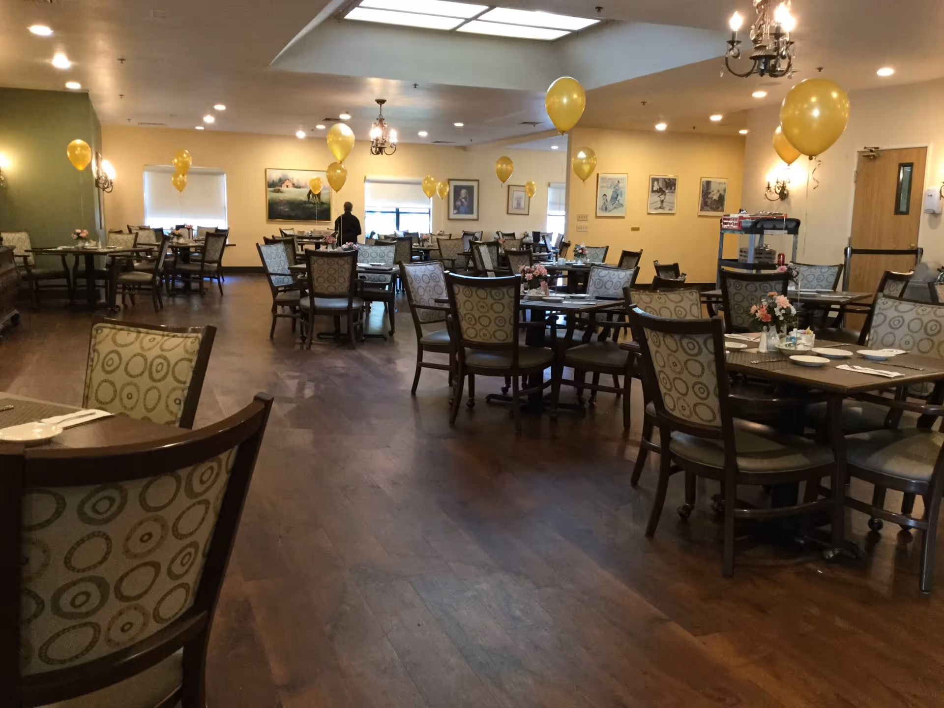 Spacious dining room with multiple tables and chairs decorated with yellow balloons and floral centerpieces.