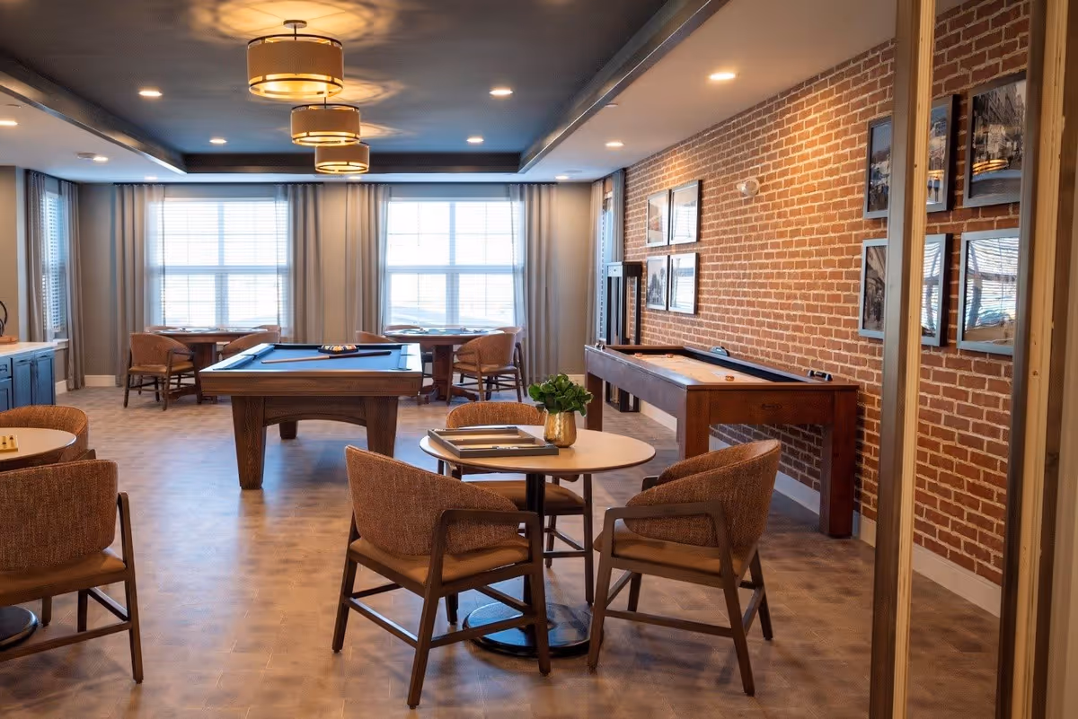 A well-lit game room with a pool table in the center, a shuffleboard table against a brick wall adorned with framed pictures, and several round tables with chairs arranged around the room. Large windows with curtains allow natural light to fill the space, and modern ceiling lights provide additional illumination.