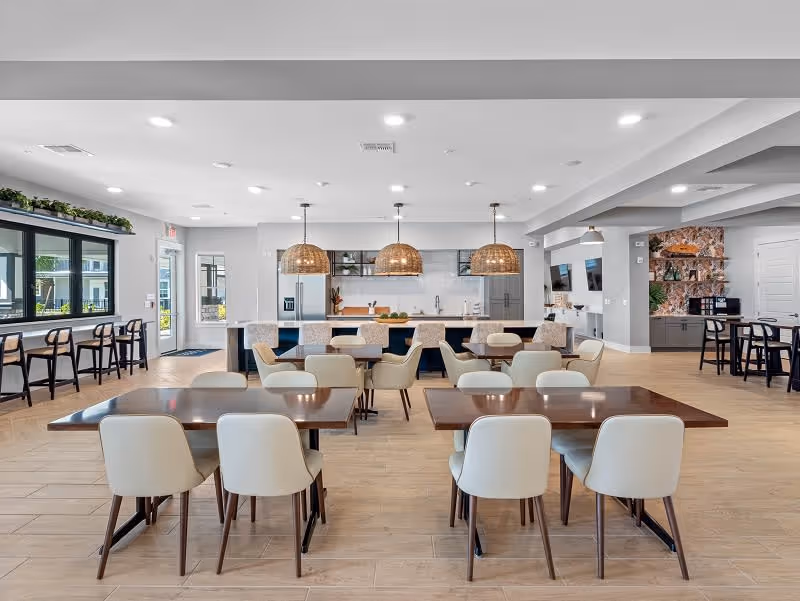 A spacious, modern dining area with multiple wooden tables and cream-colored chairs. The room features a large kitchen island with bar stools, three woven pendant lights hanging above it, and a kitchen area with stainless steel appliances in the background. Large windows on the left side let in natural light, and there are additional seating areas and decorative elements visible throughout the space.