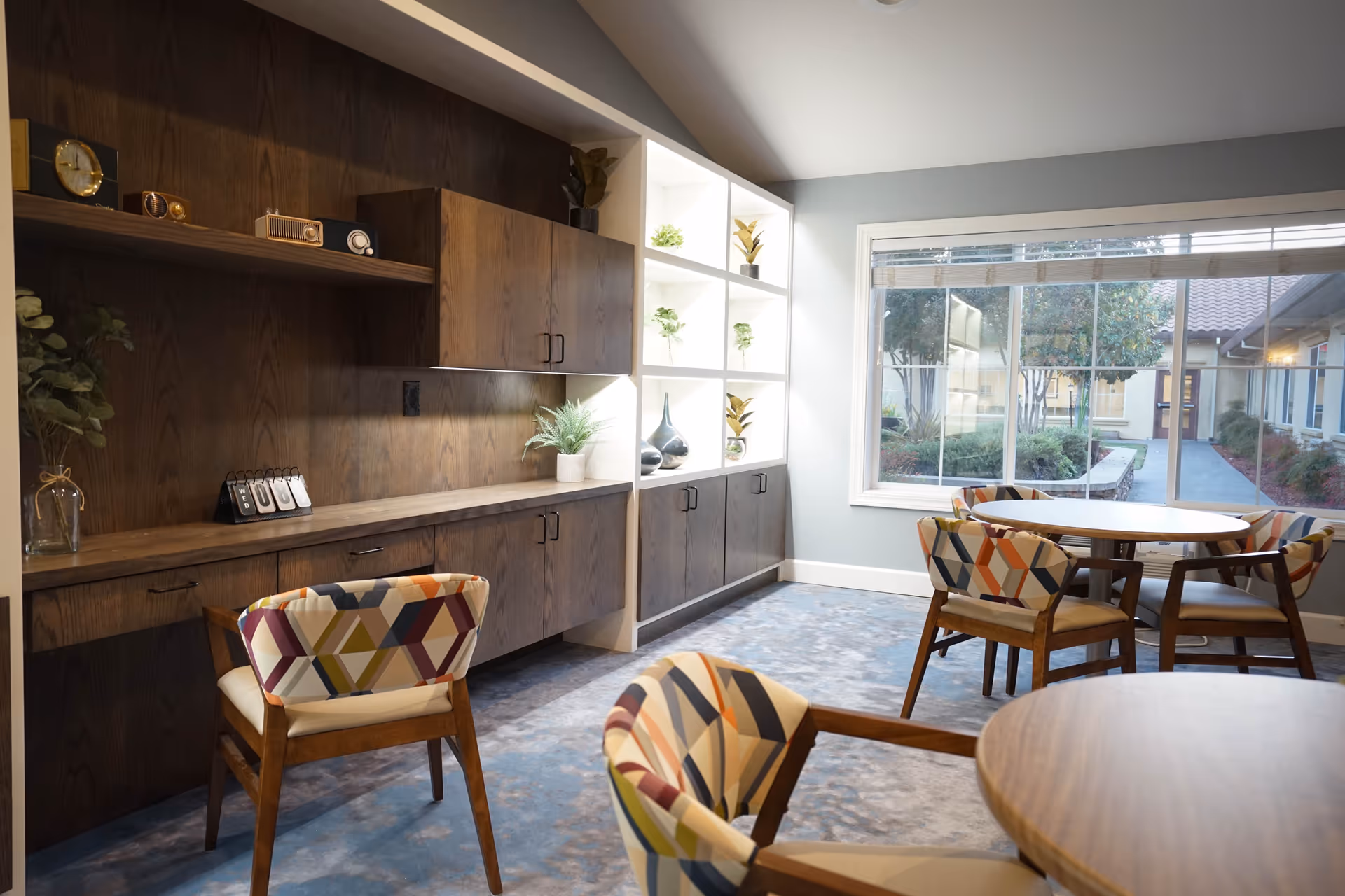 Bright common room with round tables, patterned chairs, and built-in wooden cabinets by a large window.