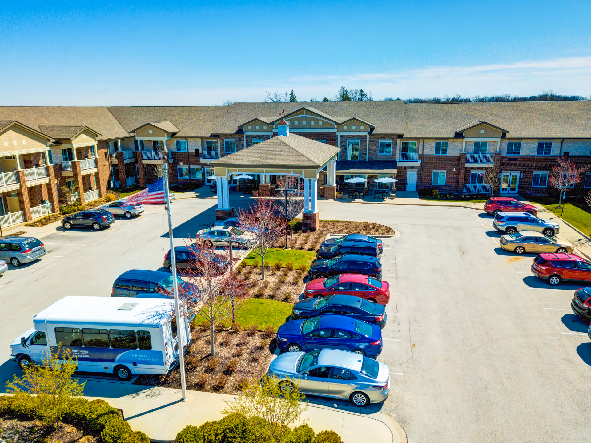 Aerial view of the Burr Ridge Senior Living building showing the covered main entrance, American flag, parking lot and multiple parked cars.