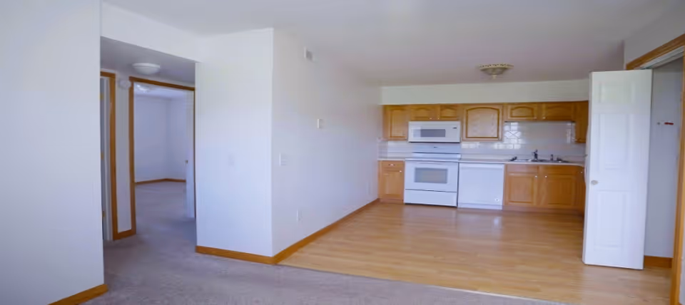Interior view of a senior living facility showing a small kitchen area with wooden cabinets, a white stove, microwave, dishwasher, and sink. The kitchen has a light wood floor, and the adjacent area is carpeted with white walls and a doorway leading to another room.