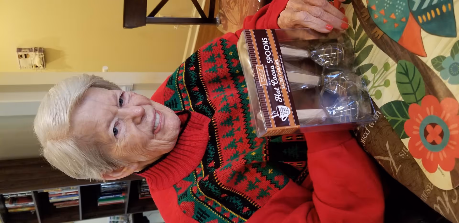 An elderly woman with short white hair wearing a red and green patterned sweater is sitting at a table with a colorful floral tablecloth. She is smiling and holding a package of hot cocoa spoons. Behind her, there is a bookshelf with books and a yellow wall.