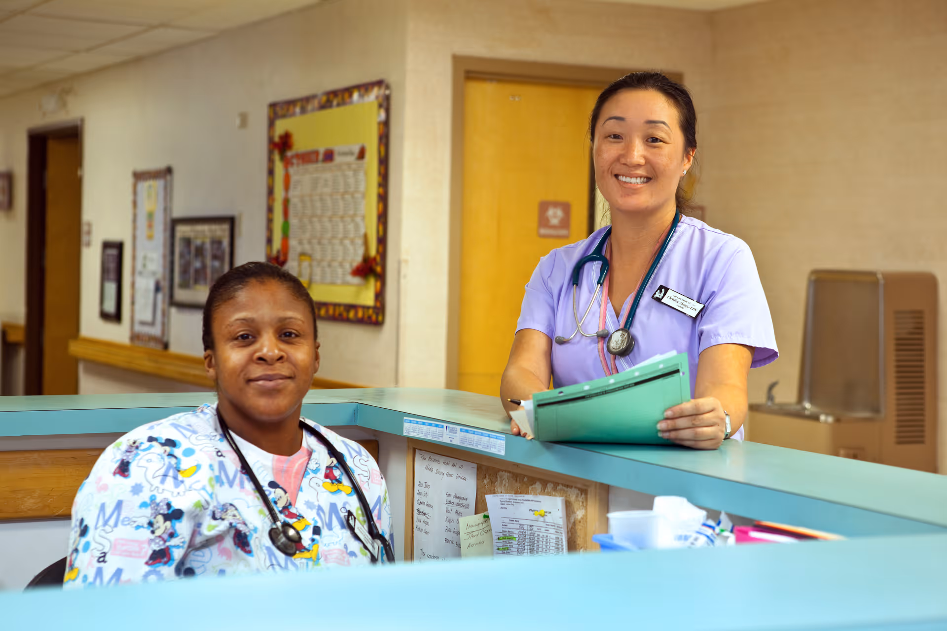 Two healthcare workers at a reception desk in a medical facility. One is seated wearing a colorful scrub top with cartoon characters and a stethoscope, and the other is standing, smiling, wearing a light purple scrub top with a stethoscope and holding a green folder. The background shows a hallway with bulletin boards and a water fountain.